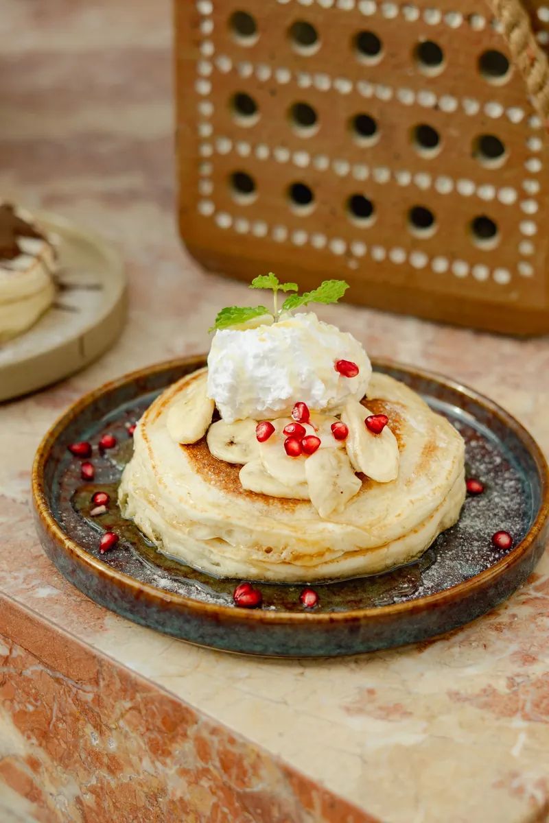 A stack of banana pancakes on a dark plate, topped with cream and pomegranate seeds.