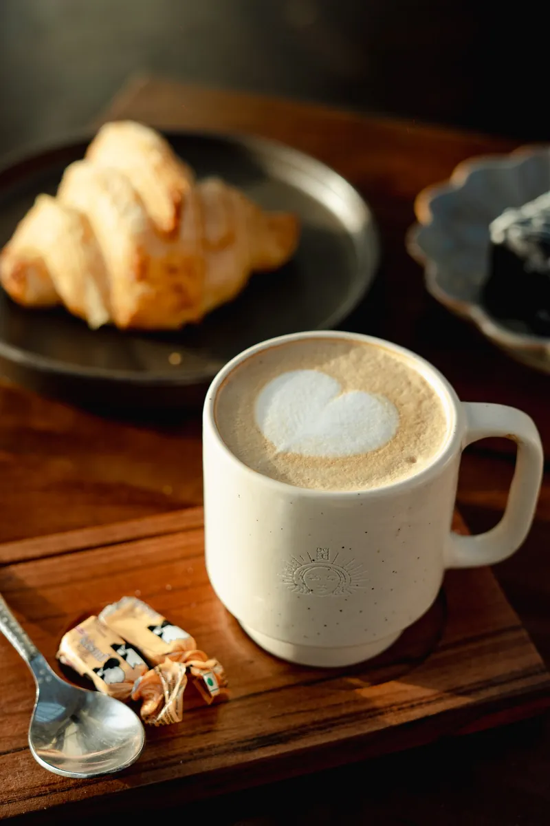 A cappuccino in a pale stoneware cup, a heart poured into the foam, a croissant behind.