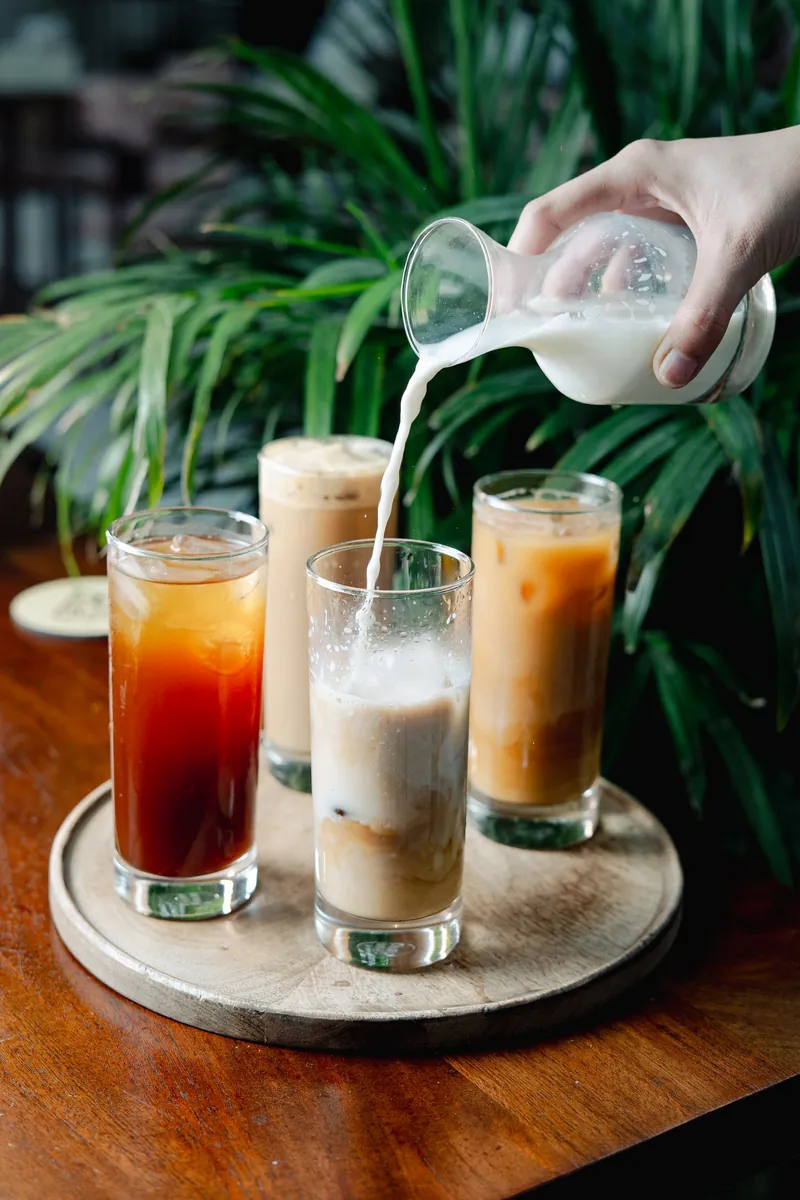 Tall glasses of cold brew and iced tea on a tray, milk being poured into one.