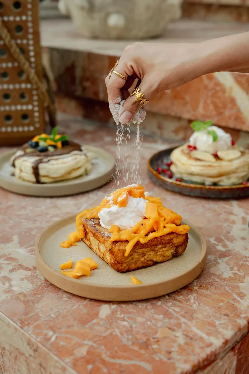 French toast under a mango sauce with a spoonful of cream, sauce being poured over it.