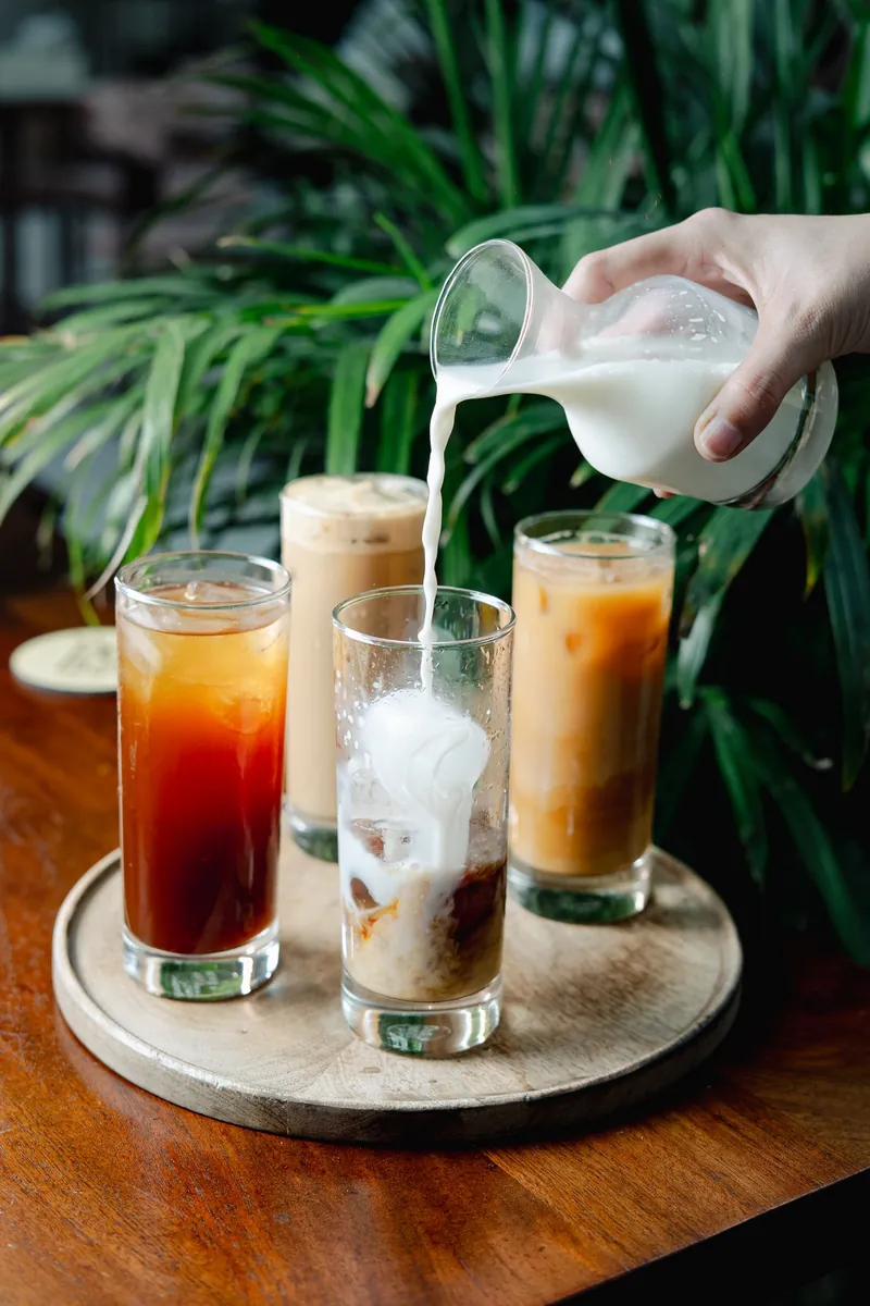Three iced coffees in tall glasses, milk being poured into one, palms behind.