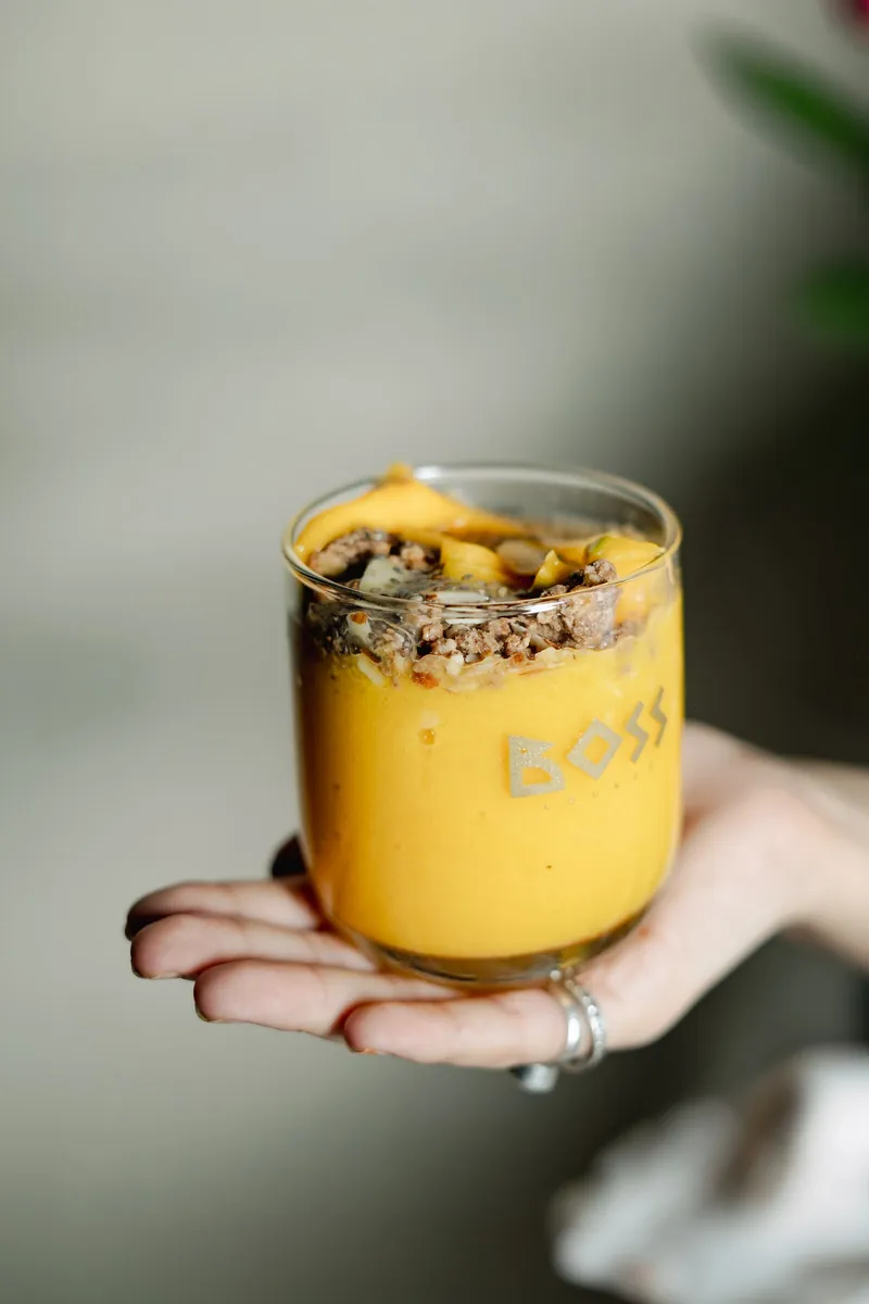 A mango, granola and yoghurt parfait in a BOSS glass, held in one hand.