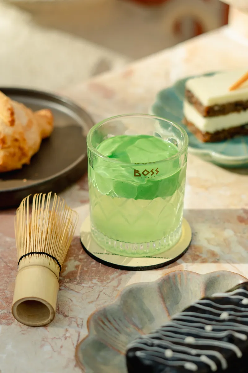 A green matcha drink in a cut BOSS tumbler beside a bamboo whisk.