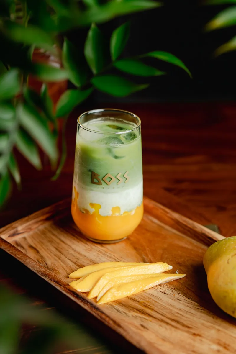 A layered cooler in a tall BOSS glass, mango at the base under cream and green, fresh mango slices beside it.