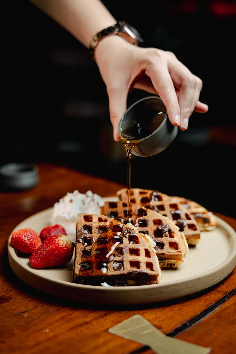 Waffles on a pale plate with strawberries, syrup being poured over them.