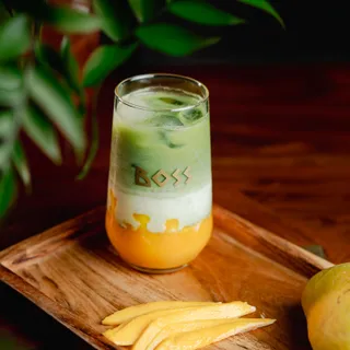 A layered cooler in a tall BOSS glass, mango at the base under cream and green, fresh mango slices beside it.