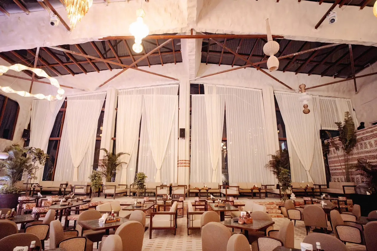 Floor-to-ceiling sheer drapes across the tall windows of the lounge, framed by palms and bulb chandeliers.