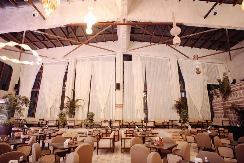 Floor-to-ceiling sheer drapes across the tall windows of the lounge, framed by palms and bulb chandeliers.