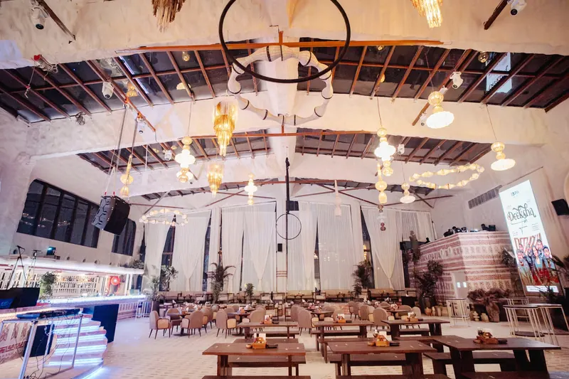 The hall seen wide, a black hoop chandelier hanging at the centre of the timber roof above the tables.