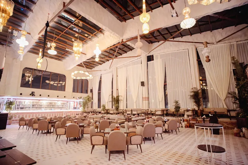 Set tables and cane chairs across the patterned stone floor of the hall, hoop chandeliers overhead.