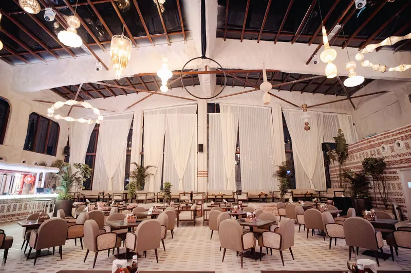 The BOSS hall from the back: rows of cane chairs and set tables under bulb chandeliers and an open timber roof, sheer drapes across the far wall.