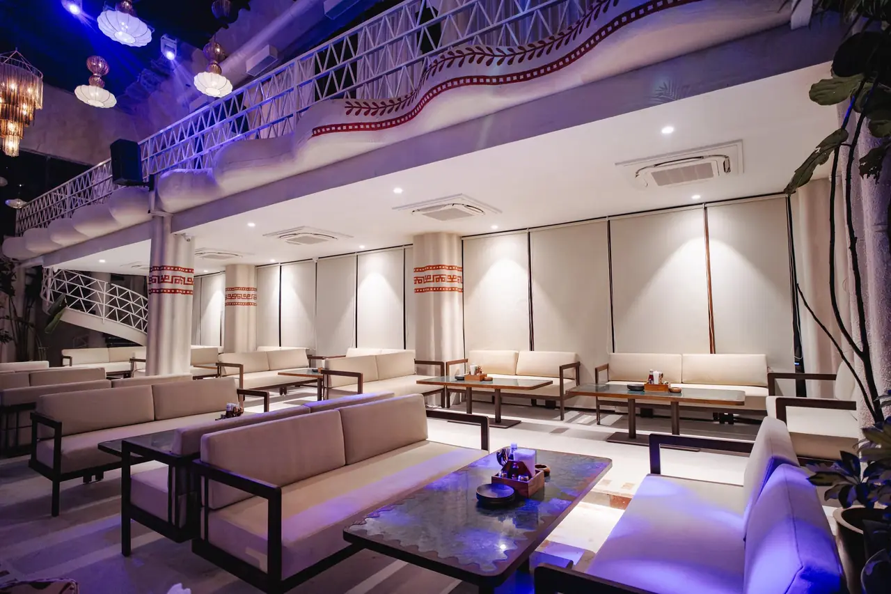 The mezzanine lounge: white sofas and low dark tables under a white lattice balcony, lit purple.