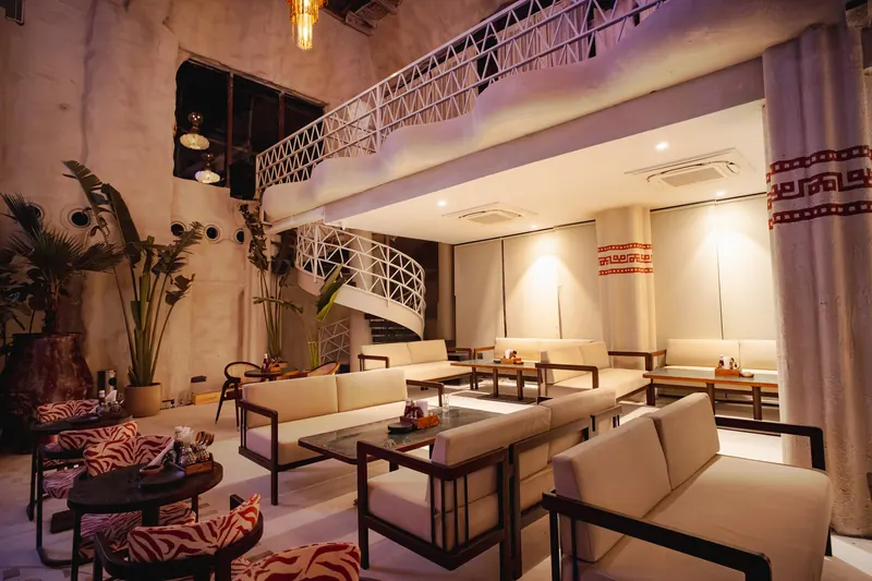 The white spiral staircase up to the mezzanine, sofas and tall plants around it in warm light.